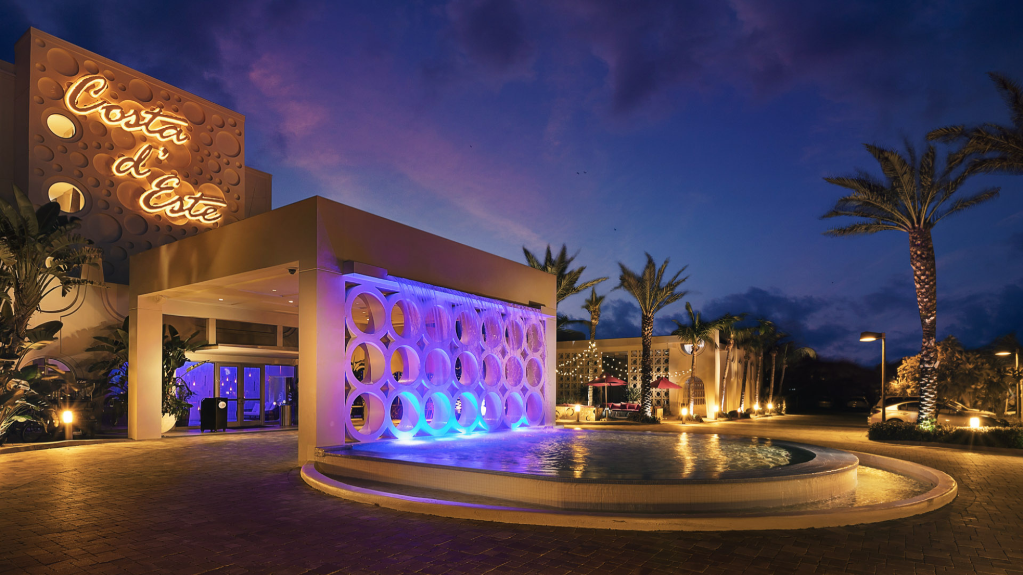 The image shows the entrance of a luxurious hotel, Costa d'Este, illuminated at night with elegant architecture and a circular water feature.