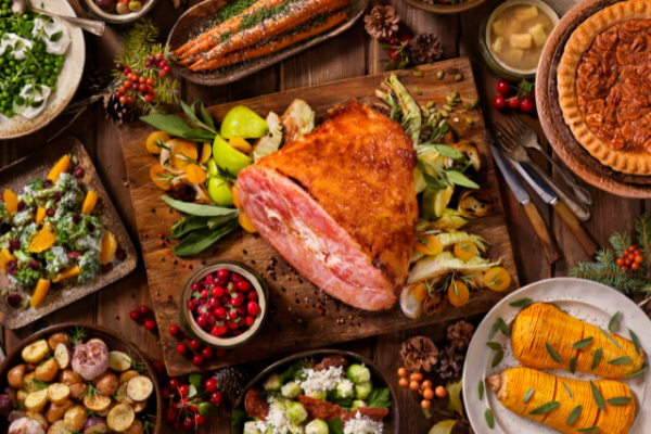 A festive spread with a glazed ham, roasted vegetables, fresh salad, pie, and garnishes, set on a rustic wooden table for a holiday feast.