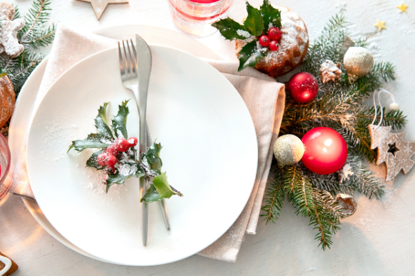 A festive table setting with a white plate, silverware, holly, candles, ornaments, and evergreen branches creates a holiday atmosphere.