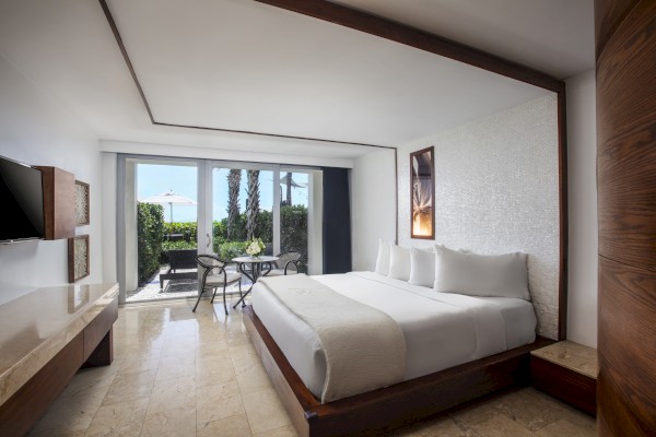 A modern hotel room with a bed, TV, and a view out to a patio with chairs and greenery beyond the sliding glass doors.