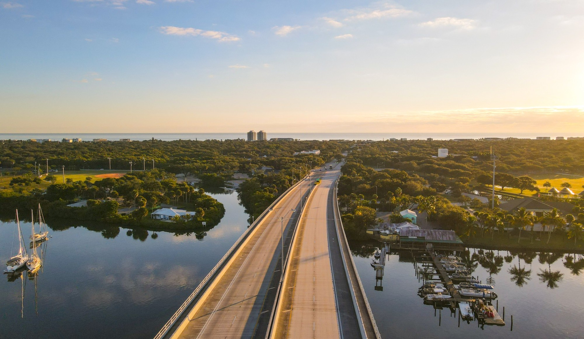 A bridge crosses over calm water surrounded by lush greenery and boats, leading toward distant buildings under a clear sky.