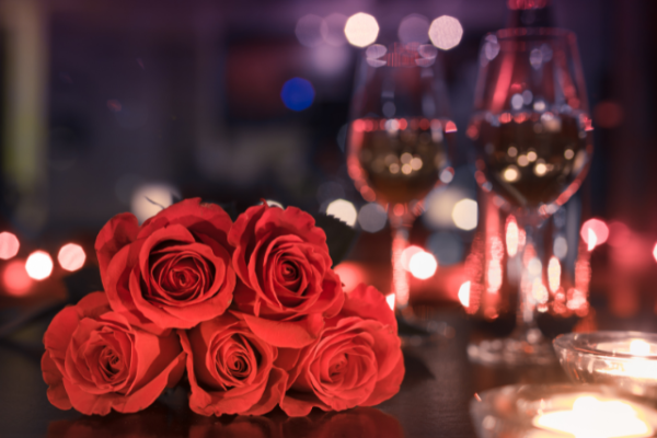 A bouquet of red roses is in the foreground with two glasses of wine and tealight candles in the background, creating a romantic ambiance.