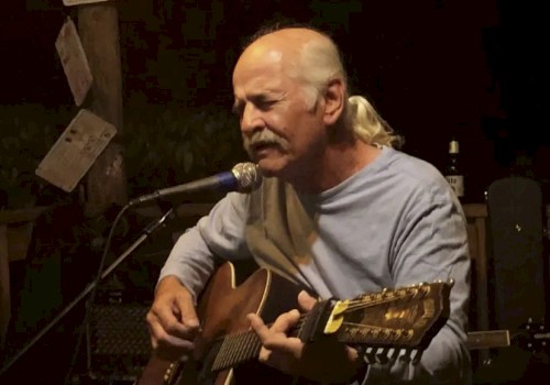 A bald man with a ponytail plays an acoustic guitar and sings into a microphone on a small stage.