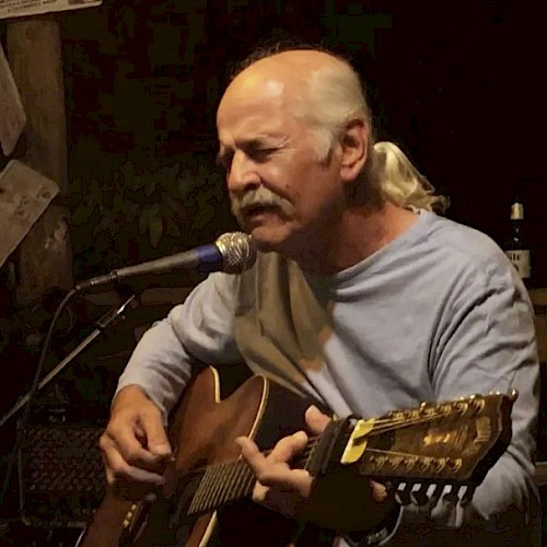 A bald man with a ponytail plays an acoustic guitar and sings into a microphone on a small stage.