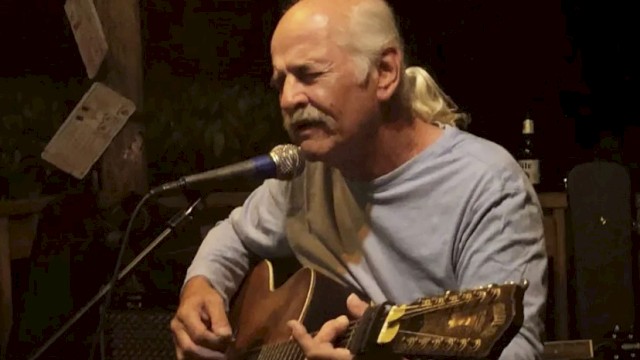 A bald man with a ponytail plays an acoustic guitar and sings into a microphone on a small stage.