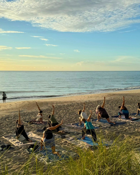 People are practicing yoga on a beach during sunrise or sunset, with a scenic ocean view in the background.