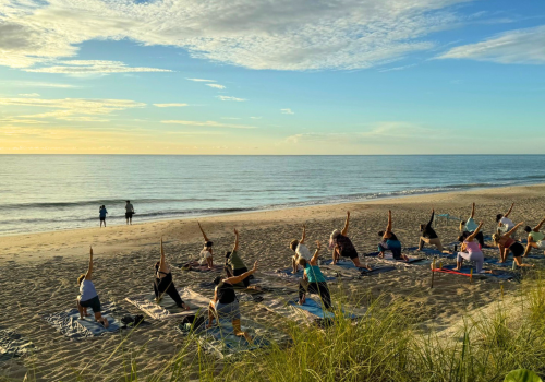 People are practicing yoga on a beach during sunrise or sunset, with a scenic ocean view in the background.