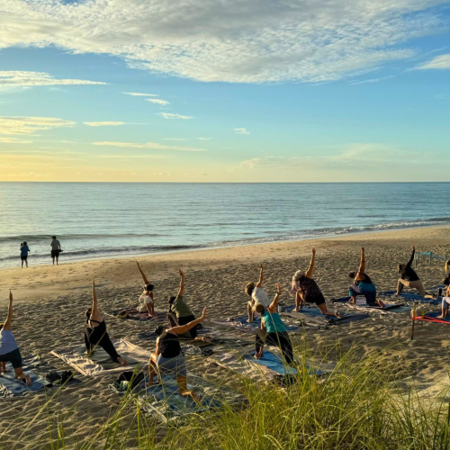 People are practicing yoga on a beach during sunrise or sunset, with a scenic ocean view in the background.