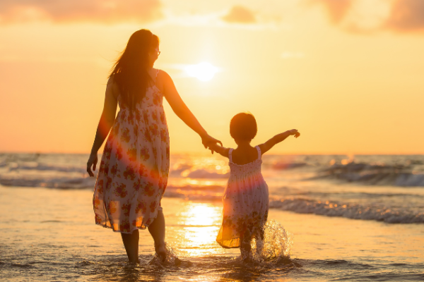 A woman and child hold hands, walking through shallow water at sunset, with the ocean waves and warm light in the background.