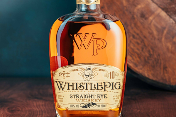 A bottle of WhistlePig 10-year Straight Rye Whiskey, labeled 100% rye, 100 proof, on a wooden surface with a barrel background.