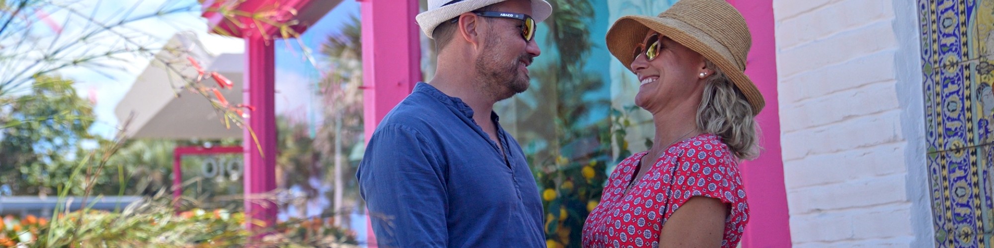 A couple, wearing hats and sunglasses, smiles at each other outside a pink-trimmed building, surrounded by greenery and a tiled wall.