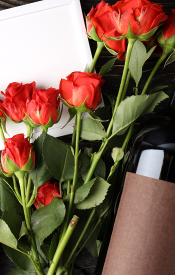 A wine bottle, red roses, a blank card, and decorative white hearts are arranged on a wooden surface, creating a romantic setting.