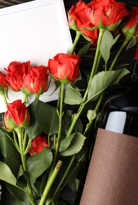 A wine bottle, red roses, a blank card, and decorative white hearts are arranged on a wooden surface, creating a romantic setting.