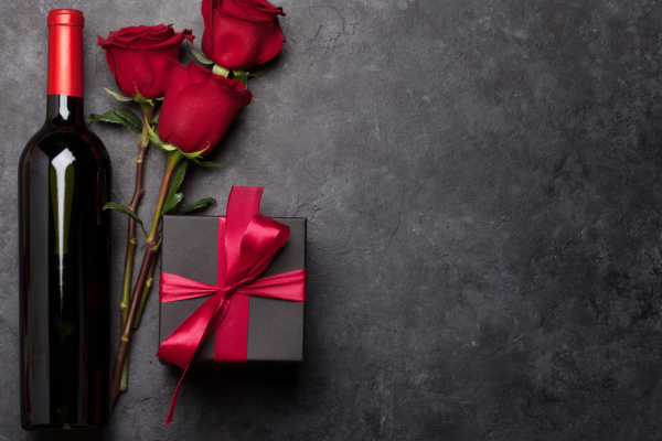 A wine bottle, three red roses, and a black gift box with a red ribbon are arranged on a dark surface.