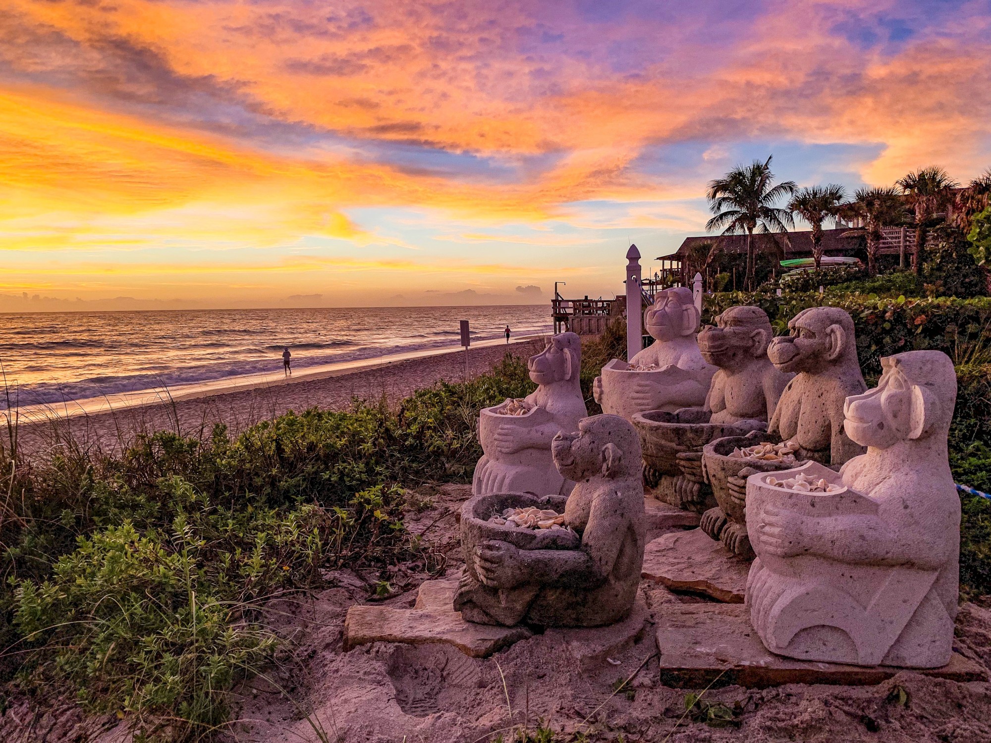 Stone monkey statues on a beach with a colorful sunset, palm trees, and ocean waves in the background.