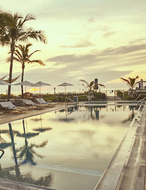 A serene poolside view with lounge chairs, umbrellas, and palm trees during a sunset, reflecting on the water's surface.