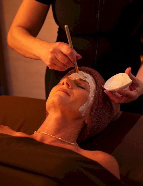A person receives a facial treatment: a therapist applies a white mask with a brush, while lying back with a head wrap. End.