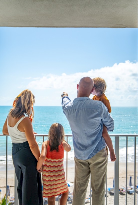 A family stands on a balcony, overlooking a sunny beach and ocean. The sky is mostly clear with a few clouds in view.