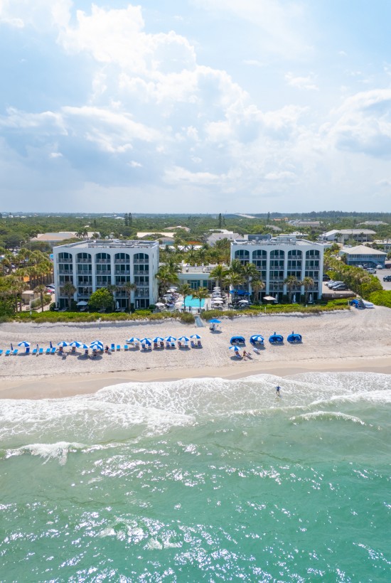 Aerial view of a beachfront resort with two white multi-story buildings, sun loungers and umbrellas on the sandy beach, and turquoise waves crashing ashore.