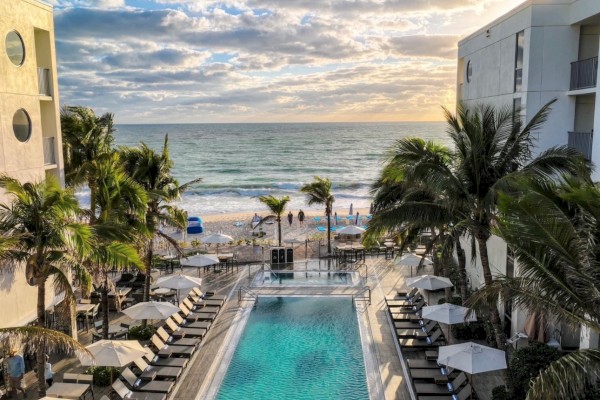 A beachfront hotel scene with a pool, loungers, umbrellas, and palm trees, overlooking the ocean under a partly cloudy sky at sunset.