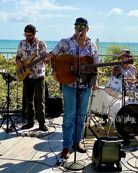 A band is performing outdoors near the ocean, featuring a keyboardist, bassist, guitarist/singer, and drummer under a sunny sky.