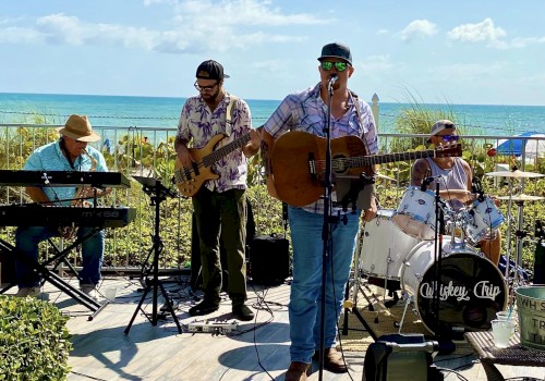 A band is performing outdoors near the ocean, featuring a keyboardist, bassist, guitarist/singer, and drummer under a sunny sky.