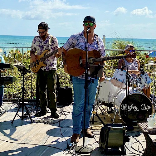 A band is performing outdoors near the ocean, featuring a keyboardist, bassist, guitarist/singer, and drummer under a sunny sky.