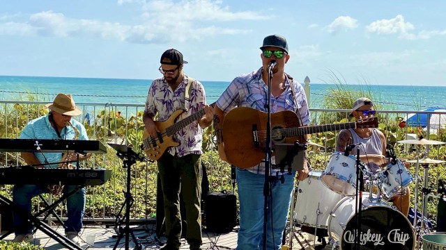A band is performing outdoors near the ocean, featuring a keyboardist, bassist, guitarist/singer, and drummer under a sunny sky.