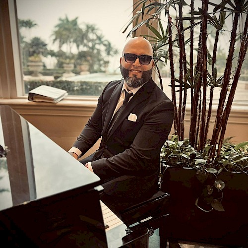 A person in a suit and sunglasses plays a grand piano indoors, with tall plants and a large window showing an outdoor scene in the background.