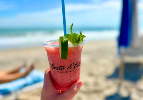 A hand holds a fruity drink with lime and mint at Costa d'Este Beach Resort & Spa, with a beach and ocean in the background.