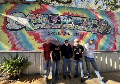 Five men stand in front of a colorful tie-dye mural with “Smoking since 1980” text, posing for a casual group photo.
