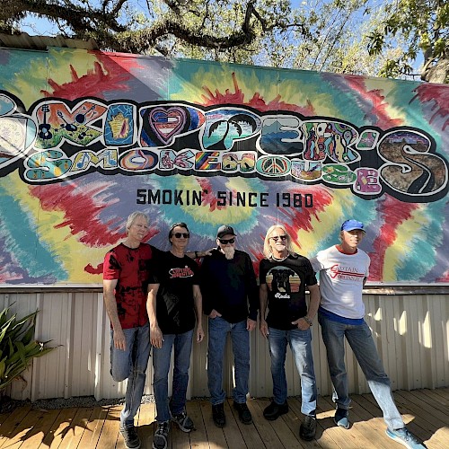 Five men stand in front of a colorful tie-dye mural with “Smoking since 1980” text, posing for a casual group photo.