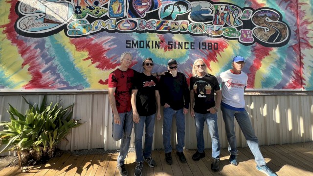 Five men stand in front of a colorful tie-dye mural with “Smoking since 1980” text, posing for a casual group photo.