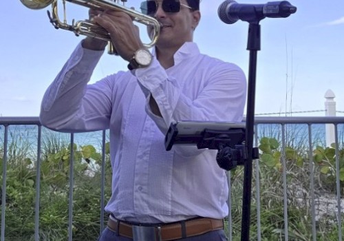 A man playing a trumpet outdoors near a fence, wearing sunglasses and a white shirt, with a microphone stand beside him against a blue sky.
