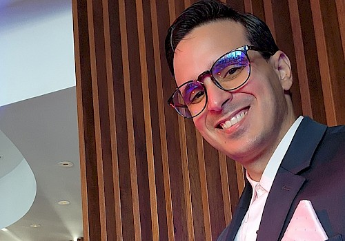 The image shows a person wearing a suit and glasses, smiling, with a wooden panel background and modern interior design.
