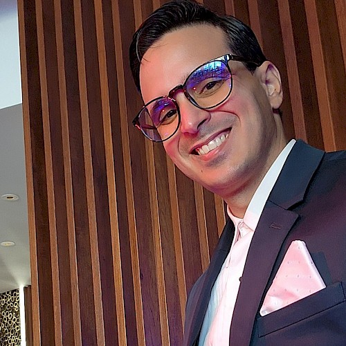 The image shows a person wearing a suit and glasses, smiling, with a wooden panel background and modern interior design.