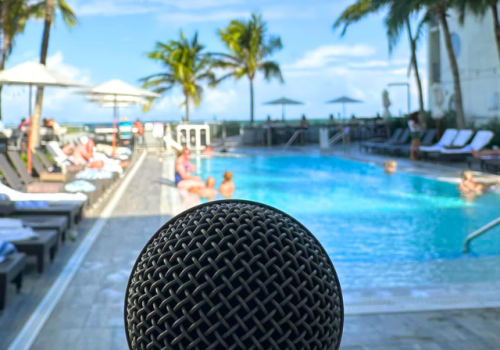 A microphone is in the foreground with a poolside setting featuring lounge chairs, people sunbathing, and palm trees in the background.