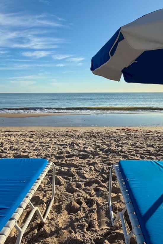 A serene beach scene with blue lounge chairs, an umbrella, and a calm sea under a clear sky. The sun reflects off the water's surface.