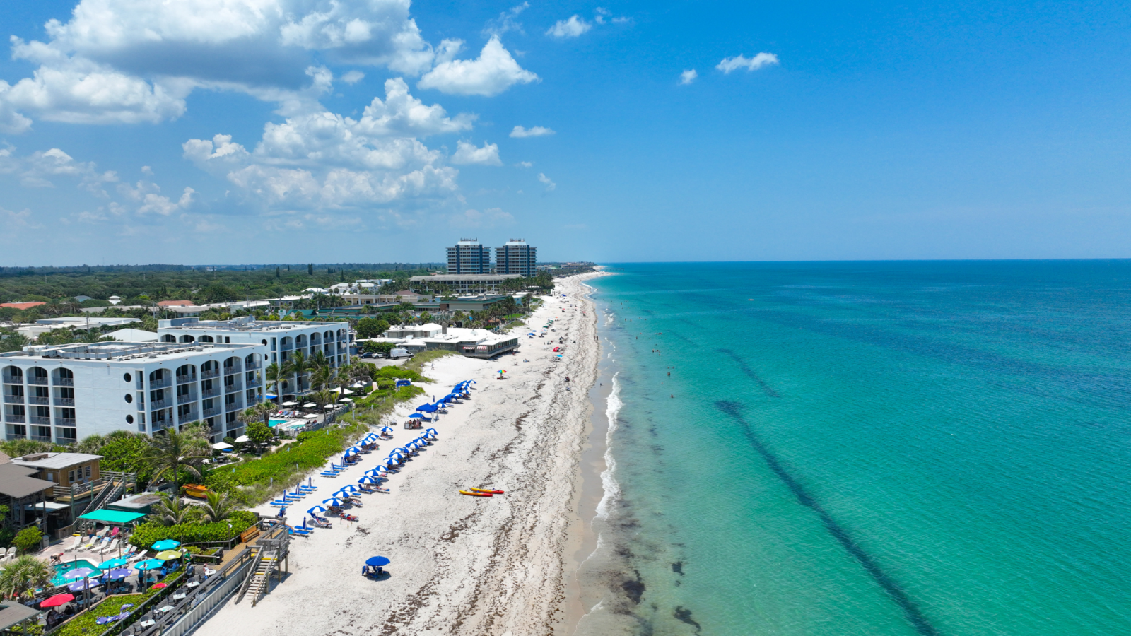 A beach scene with white sand, blue umbrellas, buildings, and turquoise water under a mostly sunny sky.