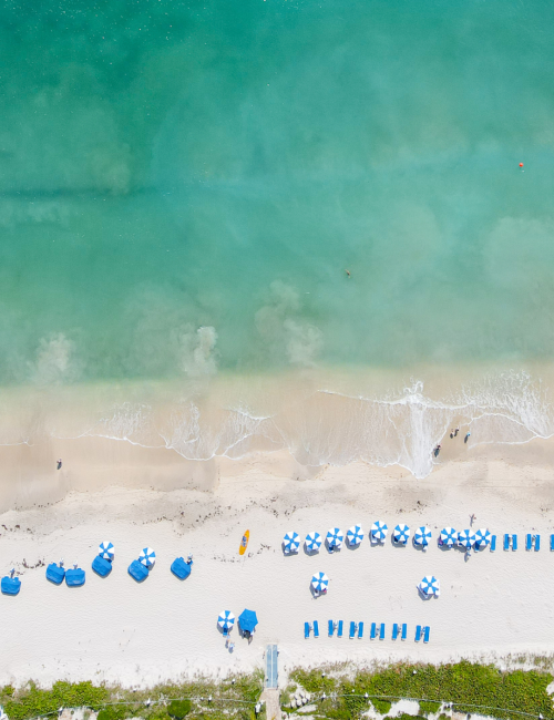 An aerial view of a beach with blue umbrellas, lounge chairs, and a swimming pool at the bottom, with clear aqua water and white sand.