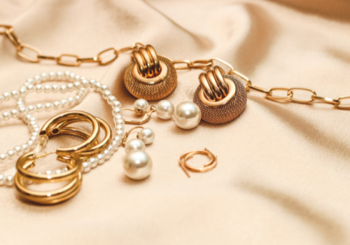 The image shows various pieces of jewelry, including pearl necklaces, gold rings, earrings, and a chain on a soft fabric surface.