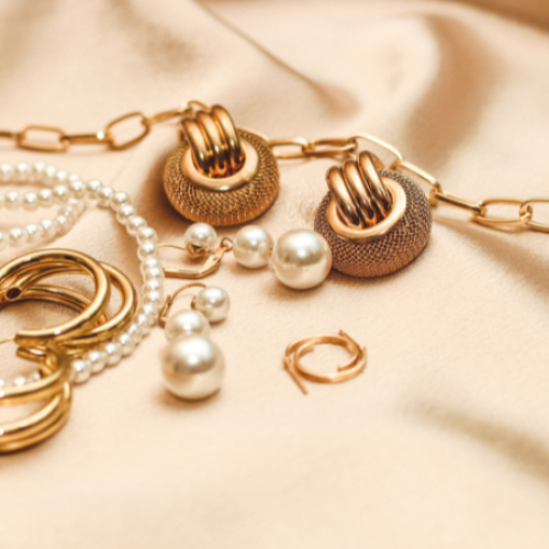 The image shows various pieces of jewelry, including pearl necklaces, gold rings, earrings, and a chain on a soft fabric surface.