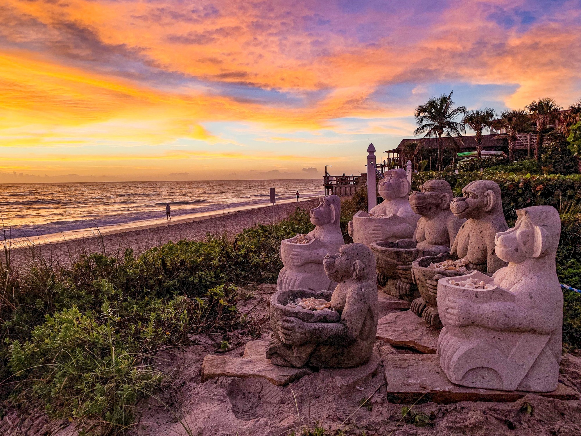 Statues of monkeys holding baskets sit by the beach at sunset, with vibrant sky and calm waves creating a serene atmosphere.