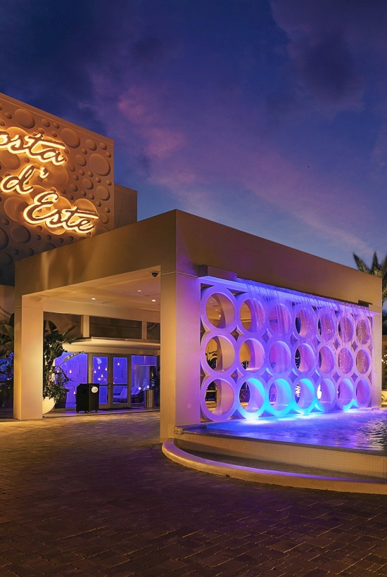 A hotel entrance at dusk features modern architecture, a circular driveway with a fountain, and illuminated palm trees in the background.