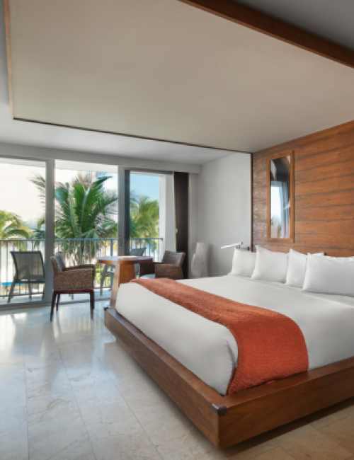 A modern hotel room with a large bed, wooden accents, a balcony with chairs, and a view of palm trees and the sky outside.