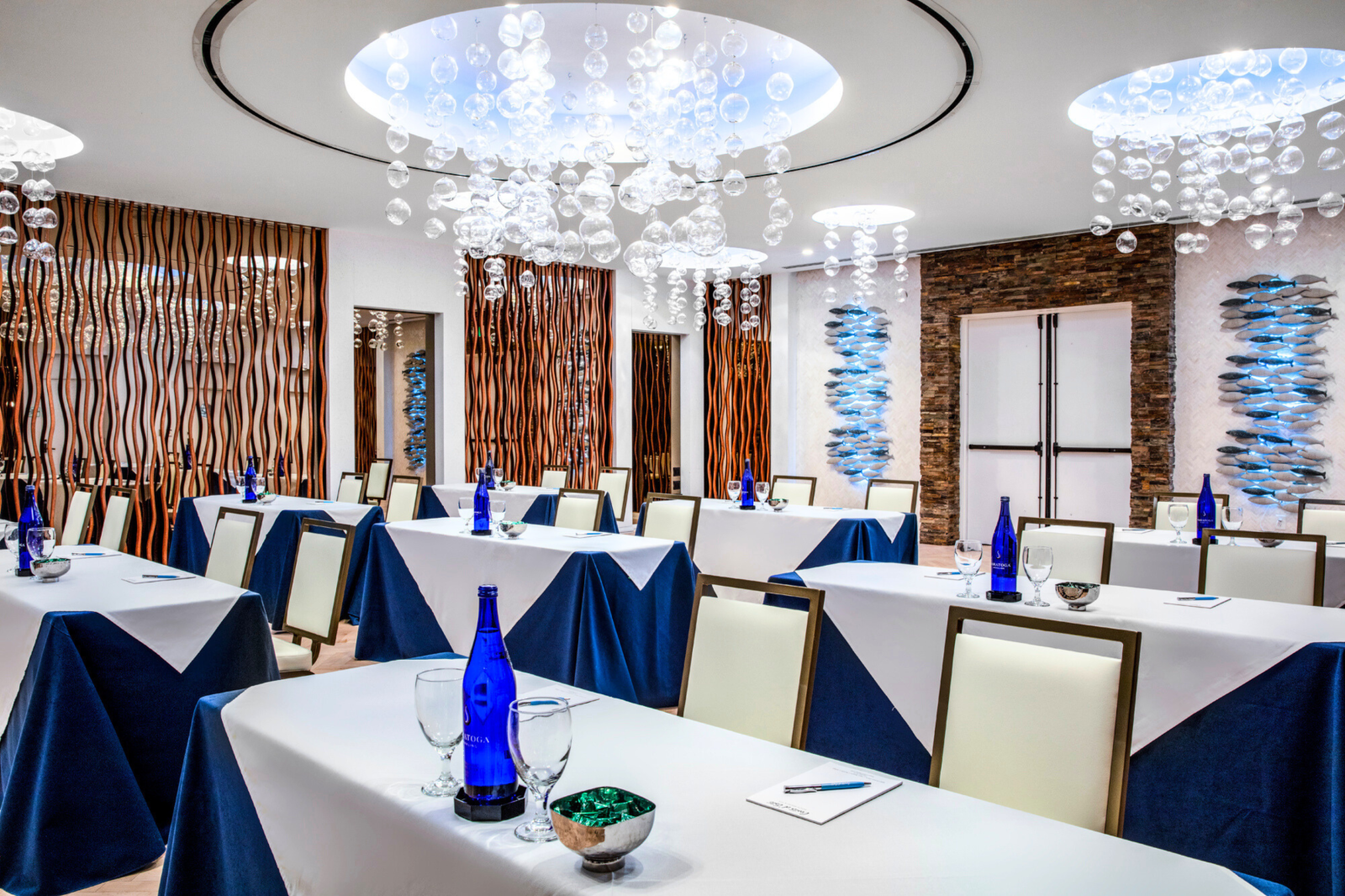 The image shows a modern conference room with tables set with blue bottles, glasses, and notebooks, featuring artistic lighting fixtures.