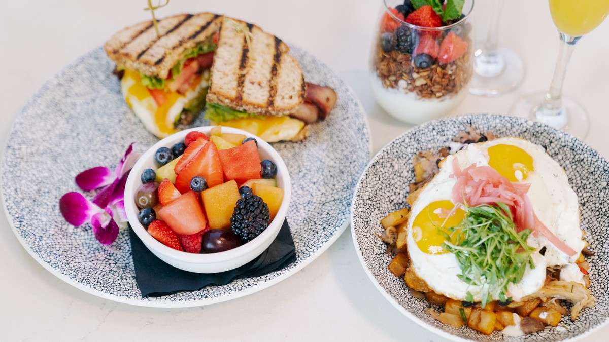 The image shows a plate with a sandwich, a bowl of mixed fruit, a dish with eggs and garnish, and parfait glasses in the background.