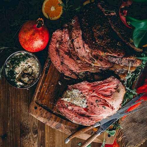 The image shows a sliced Prime Rib on a wooden board with herbs, a knife, orange slices, a pomegranate, and a bowl of seasoning.