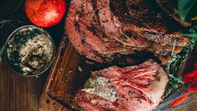 The image shows a sliced Prime Rib on a wooden board with herbs, a knife, orange slices, a pomegranate, and a bowl of seasoning.