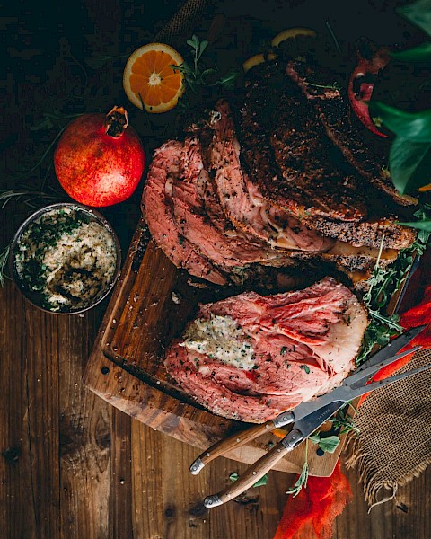 The image shows a sliced Prime Rib on a wooden board with herbs, a knife, orange slices, a pomegranate, and a bowl of seasoning.